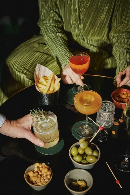 Hands reaching across a low table of cocktails, olives and almonds